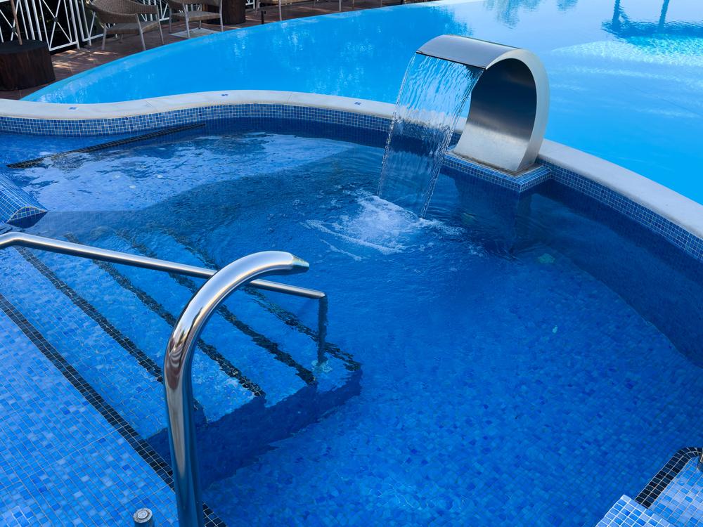 Resurfaced swimming pool with blue tile, spa area, stainless steel handrail, and waterfall feature illustrating the cost to resurface a pool.